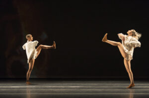 Two dancers in white facing each other, with their legs kicked up high.