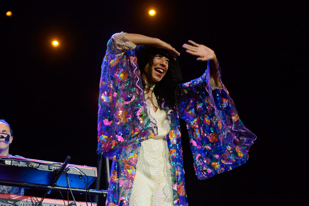 A woman with curly hair sings into a microphone on stage, illuminated by multiple spotlights. Another woman in a blue dress stands in the background, also singing.