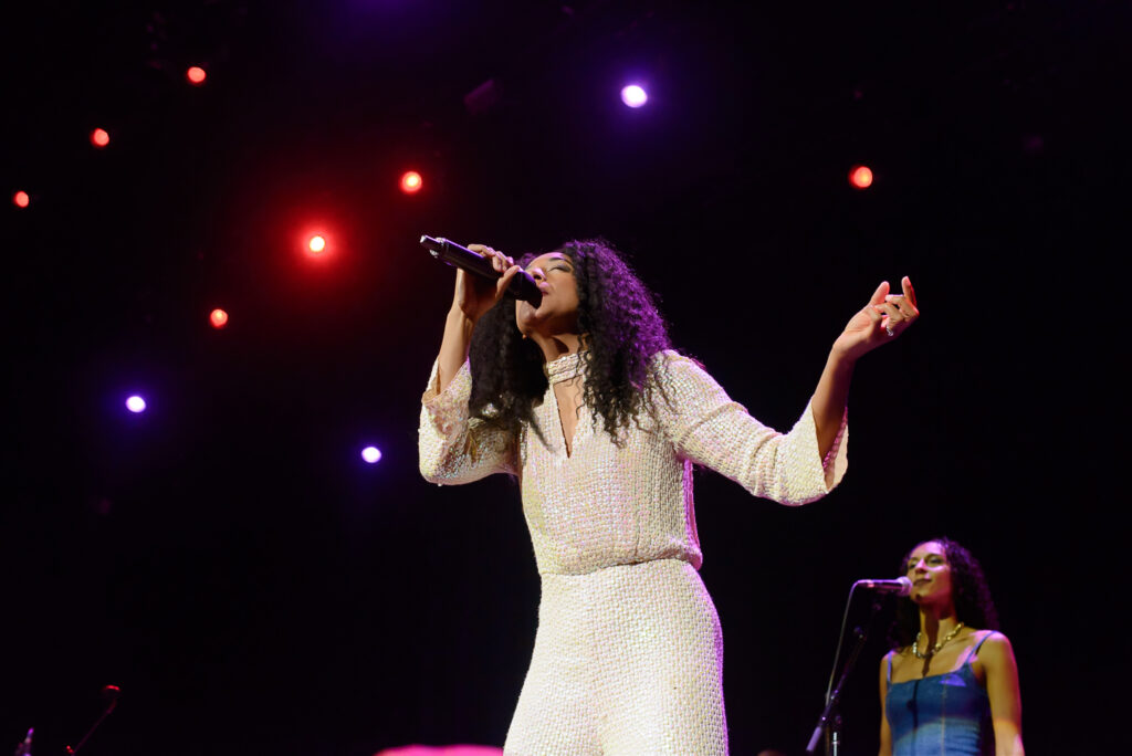 A woman with curly hair sings into a microphone on stage, illuminated by multiple spotlights. Another woman in a blue dress stands in the background, also singing.