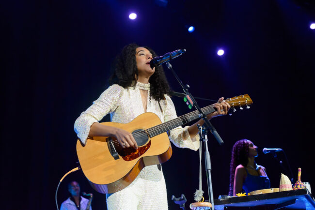 A woman with curly hair sings into a microphone on stage, illuminated by multiple spotlights. Another woman in a blue dress stands in the background, also singing.