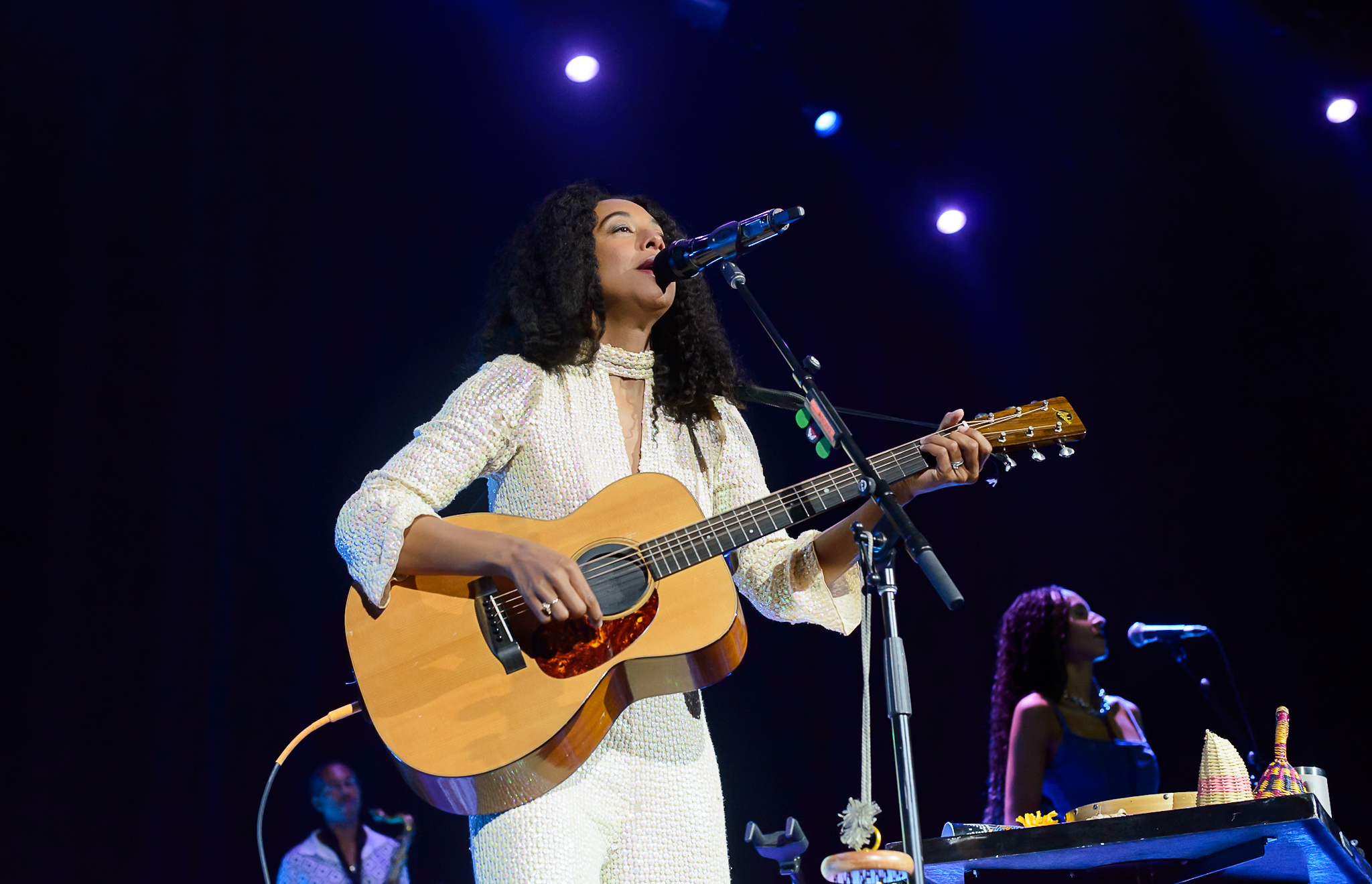 A woman with curly hair sings into a microphone on stage, illuminated by multiple spotlights. Another woman in a blue dress stands in the background, also singing.