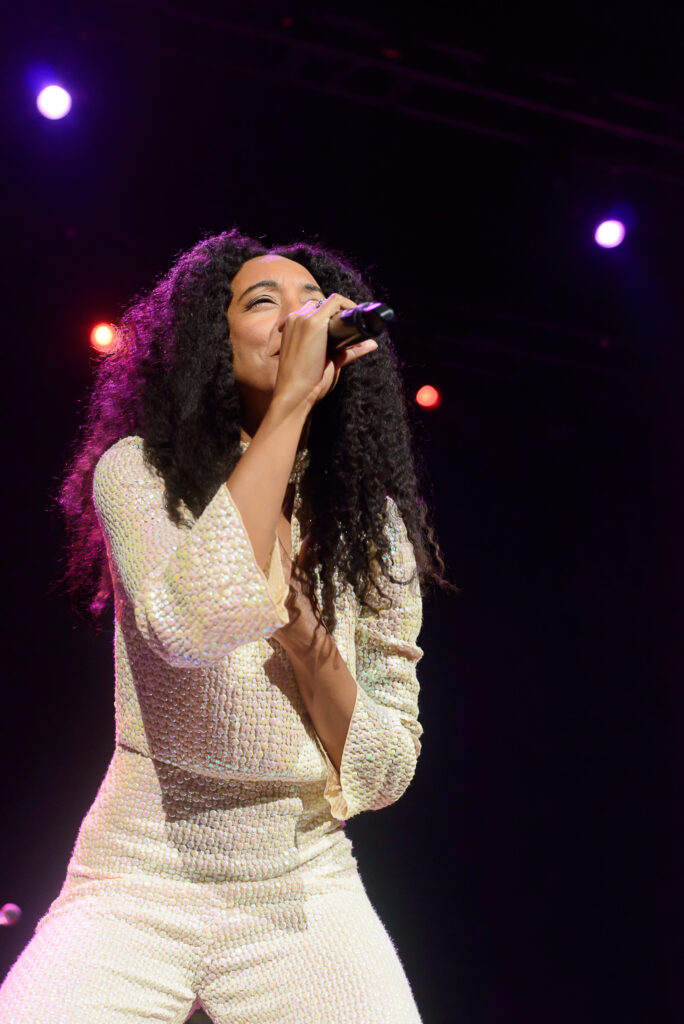 A woman with curly hair sings into a microphone on stage, illuminated by multiple spotlights. Another woman in a blue dress stands in the background, also singing.